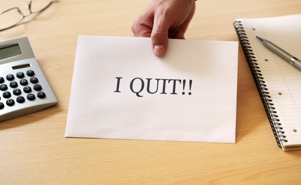 Hand holding "I QUIT!!" paper over desk with calculator and notebook